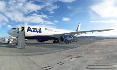 A imagem mostra um avião da companhia aérea Azul estacionado na pista de um aeroporto em um dia claro, com céu azul e algumas nuvens.