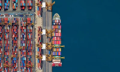 Vista aérea de um porto com contêineres coloridos organizados à esquerda, guindastes amarelos no centro-esquerdo, um navio cargueiro atracado à direita e o mar azul profundo ao fundo.