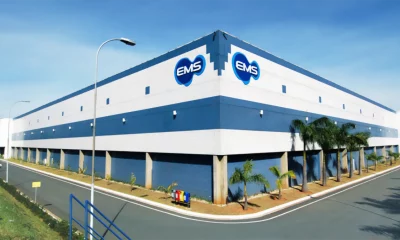 Fachada de um grande edifício industrial nas cores branco e azul com logos azuis da "EMS", palmeiras ao longo da calçada e céu azul ao fundo.