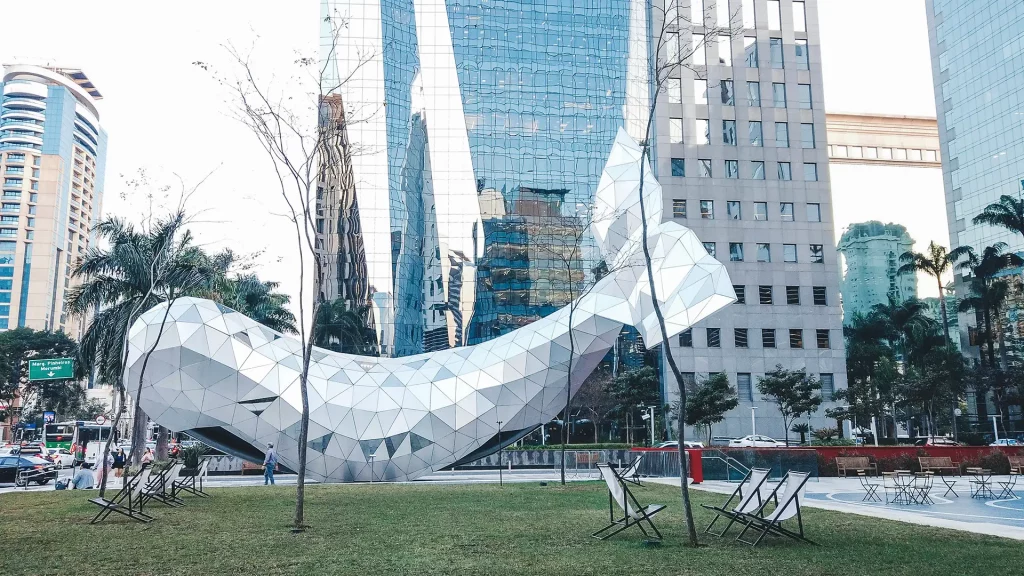 A imagem exibe uma famosa escultura futurista em formato de baleira localizada na Avenida Brigadeiro Faria Lima, na cidade de São Paulo (SP). Observam-se arranha-céus de vidro ao fundo e a escultura sobre um gramado com cadeiras de praia e algumas árvores sem folhas, sugerindo um espaço público de relaxamento.