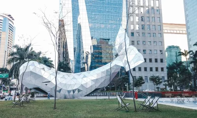 A imagem exibe uma famosa escultura futurista em formato de baleira localizada na Avenida Brigadeiro Faria Lima, na cidade de São Paulo (SP). Observam-se arranha-céus de vidro ao fundo e a escultura sobre um gramado com cadeiras de praia e algumas árvores sem folhas, sugerindo um espaço público de relaxamento.