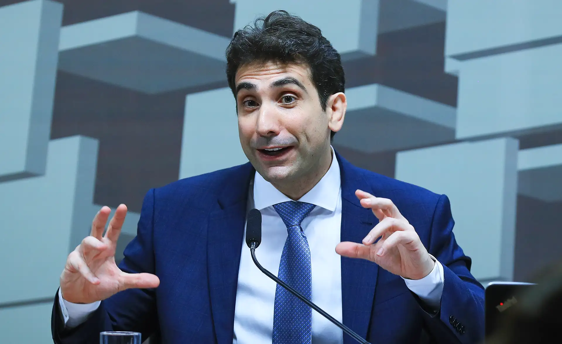 Portrait of economist Gabriel Galípolo, a man with dark hair, wearing a dark blue suit, white shirt and light blue tie, gesturing while speaking into a microphone with a background of gray geometric figures.