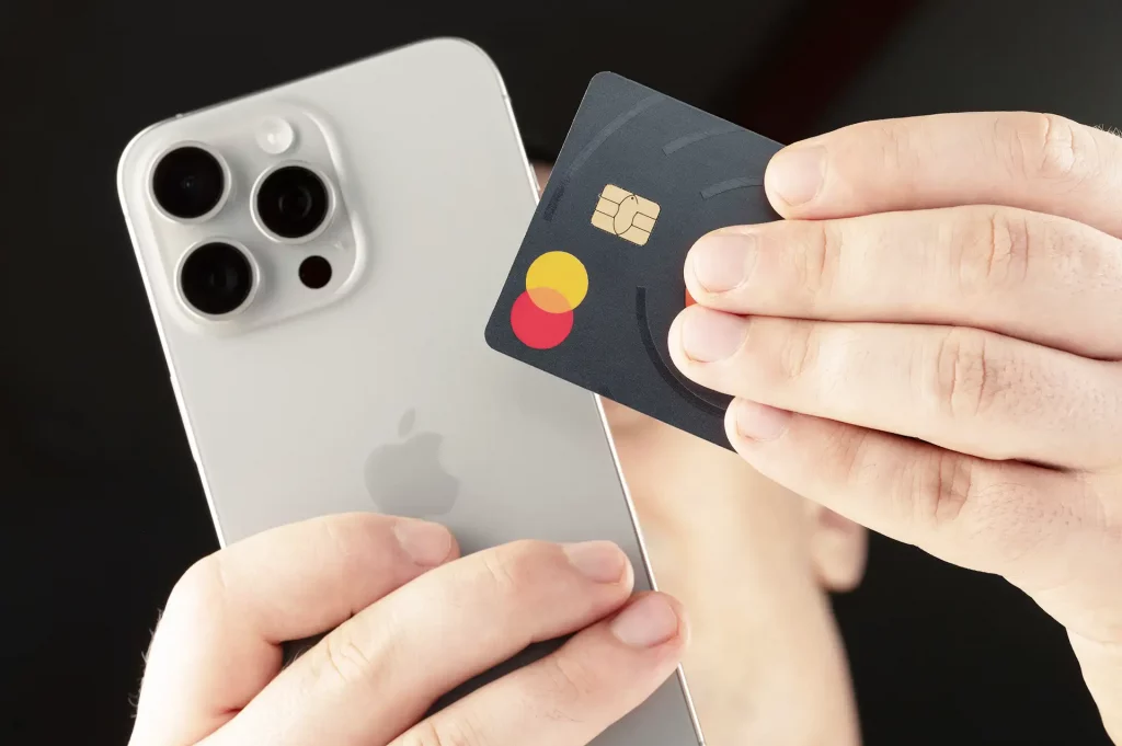 Mãos segurando um iPhone prateado e um cartão Mastercard de design escuro e minimalista.