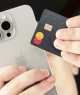 Mãos segurando um iPhone prateado e um cartão Mastercard de design escuro e minimalista.