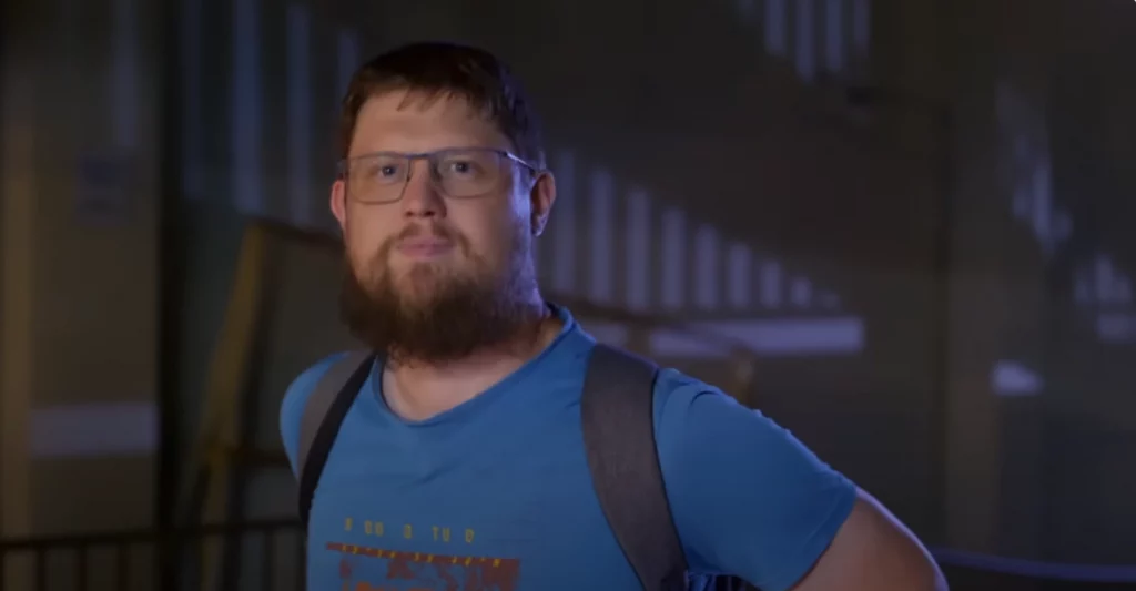 Frame do trailer do documentário “Money Electric: The Bitcoin Mystery”, onde se vê o O desenvolvedor de software Peter Todd, um Homem barbudo com óculos, camiseta azul e mochila preta em ambiente interno.