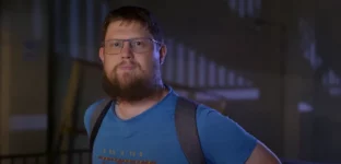 Frame do trailer do documentário “Money Electric: The Bitcoin Mystery”, onde se vê o O desenvolvedor de software Peter Todd, um Homem barbudo com óculos, camiseta azul e mochila preta em ambiente interno.