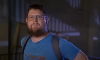 Frame do trailer do documentário “Money Electric: The Bitcoin Mystery”, onde se vê o O desenvolvedor de software Peter Todd, um Homem barbudo com óculos, camiseta azul e mochila preta em ambiente interno.