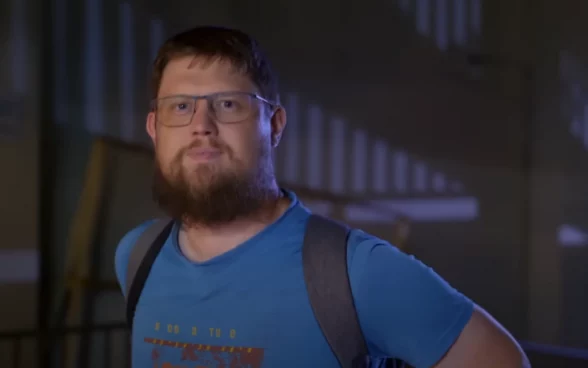 Frame do trailer do documentário “Money Electric: The Bitcoin Mystery”, onde se vê o O desenvolvedor de software Peter Todd, um Homem barbudo com óculos, camiseta azul e mochila preta em ambiente interno.