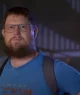 Frame do trailer do documentário “Money Electric: The Bitcoin Mystery”, onde se vê o O desenvolvedor de software Peter Todd, um Homem barbudo com óculos, camiseta azul e mochila preta em ambiente interno.