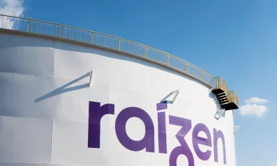 Tanque industrial branco com logotipo "raizen" em roxo, escada lateral e céu azul claro ao fundo.