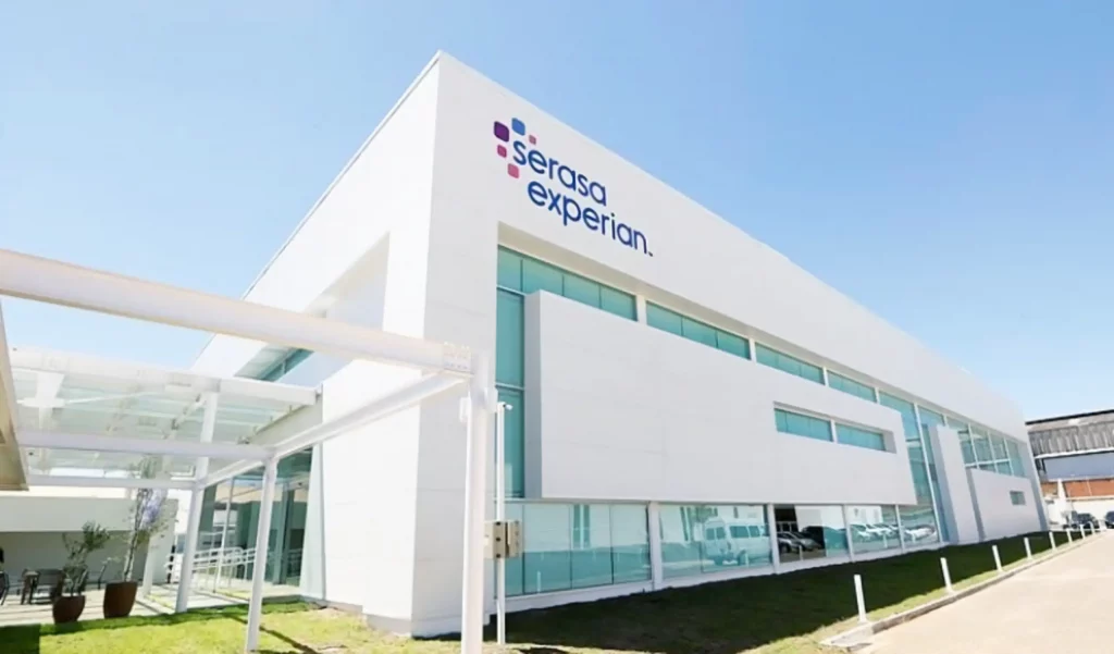 Fachada de prédio com letreiro da empresa Serasa Experian. O prédio tem acabamento em alvenaria branca e vidros azuis espelhados. A imagem tem uma perspectiva forçada, onde se vê a frente a lateral esquerda da edificação.