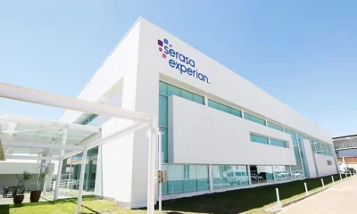 Fachada de prédio com letreiro da empresa Serasa Experian. O prédio tem acabamento em alvenaria branca e vidros azuis espelhados. A imagem tem uma perspectiva forçada, onde se vê a frente a lateral esquerda da edificação.