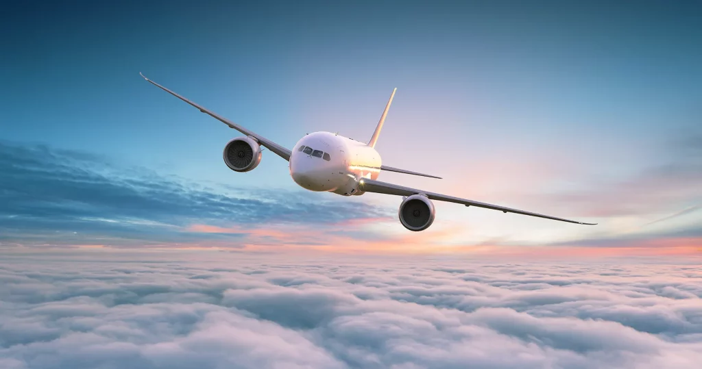 A imagem mostra um avião branco voando inclinado acima de um mar de nuvens com um céu de tons azul, rosa e laranja ao fundo.