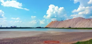 Uma paisagem industrial com uma pilha de sal, uma máquina de mineração, uma lagoa azul esverdeada, terreno arenoso e uma fábrica ao fundo, sob um céu parcialmente nublado.