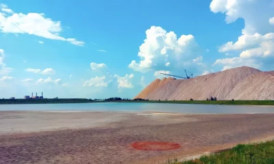 Uma paisagem industrial com uma pilha de sal, uma máquina de mineração, uma lagoa azul esverdeada, terreno arenoso e uma fábrica ao fundo, sob um céu parcialmente nublado.