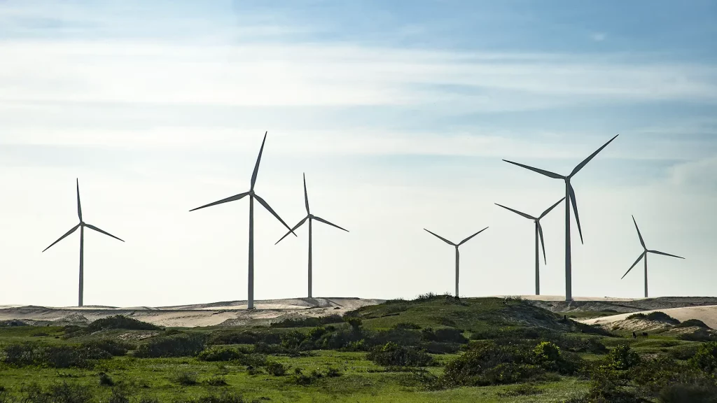 Campo de turbinas eólicas em paisagem costeira com vegetação rasteira e céu parcialmente nublado.