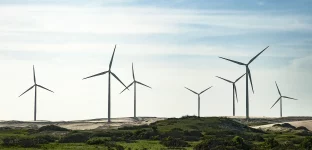 Campo de turbinas eólicas em paisagem costeira com vegetação rasteira e céu parcialmente nublado.