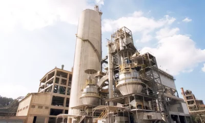 Uma instalação industrial complexa com silos e torres sob um céu azul, cercada por algumas árvores e uma chaminé.