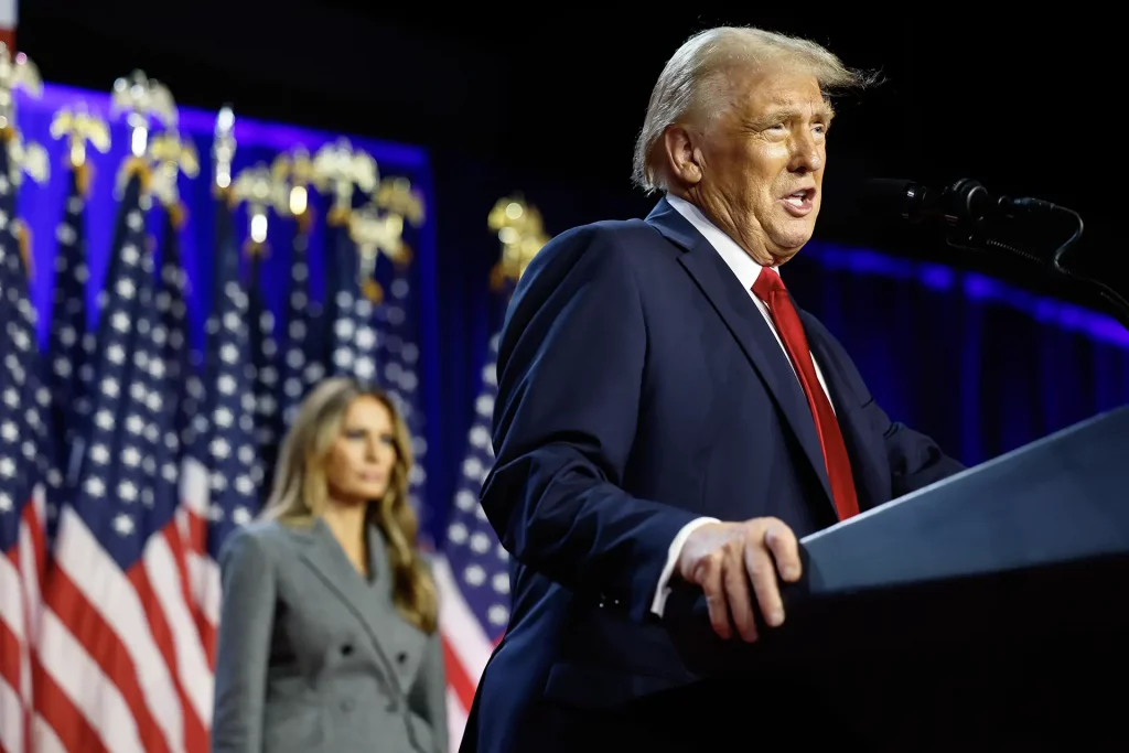 O presidente eleito dos EUA, Donald Trump, um homem em terno azul escuro discursando em um palco com bandeiras americanas ao fundo, observado por sua esposa, Melania Trump, uma mulher de casaco cinza.