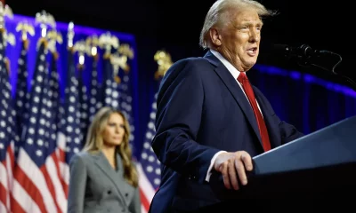 O presidente eleito dos EUA, Donald Trump, um homem em terno azul escuro discursando em um palco com bandeiras americanas ao fundo, observado por sua esposa, Melania Trump, uma mulher de casaco cinza.