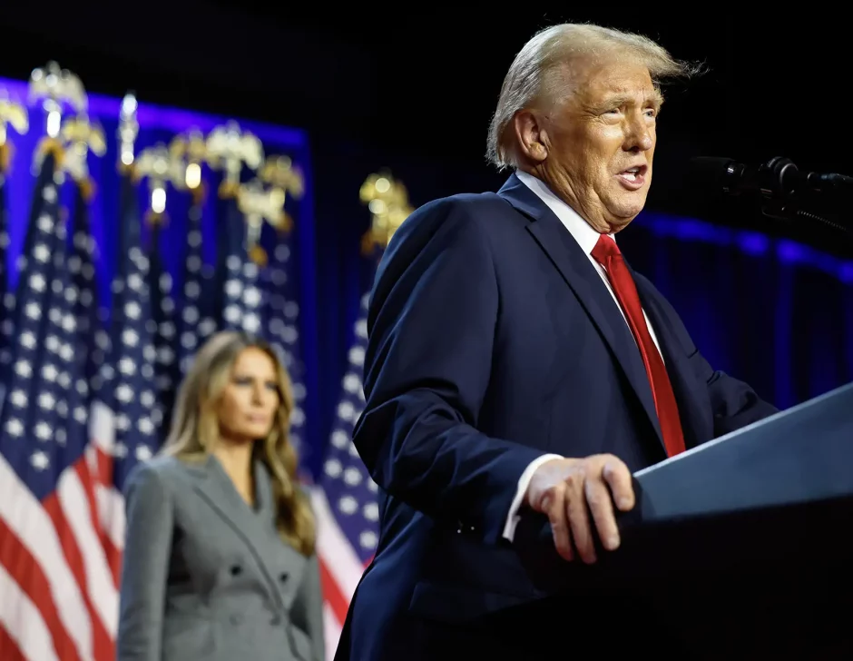 O presidente eleito dos EUA, Donald Trump, um homem em terno azul escuro discursando em um palco com bandeiras americanas ao fundo, observado por sua esposa, Melania Trump, uma mulher de casaco cinza.