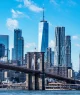 A imagem retrata o skyline de Nova York com a Ponte do Brooklyn em primeiro plano e Manhattan ao fundo, destacando o One World Trade Center contra um céu azul com nuvens esparsas.