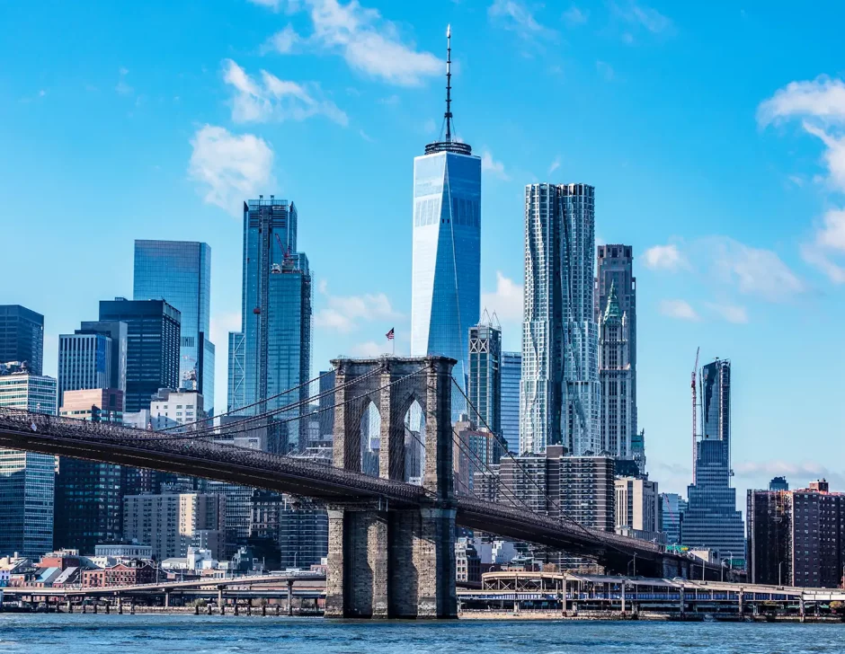 A imagem retrata o skyline de Nova York com a Ponte do Brooklyn em primeiro plano e Manhattan ao fundo, destacando o One World Trade Center contra um céu azul com nuvens esparsas.