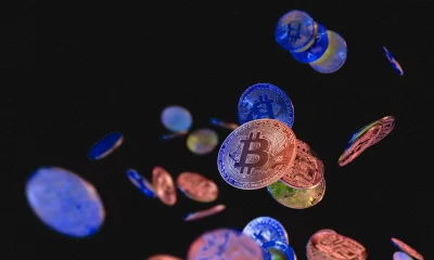 Moedas virtuais flutuam contra um fundo preto, com um Bitcoin destacado no centro, rodeado por tons metálicos de azul, vermelho e dourado.