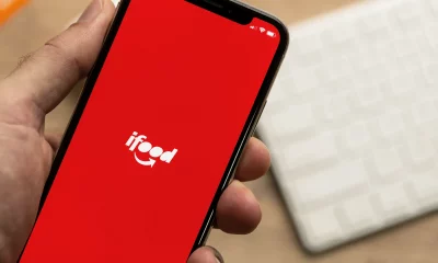 Uma mão segura um smartphone exibindo o aplicativo iFood em uma tela vermelha, com o logotipo branco em destaque e um teclado branco desfocado ao fundo.