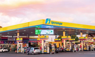 Posto de combustíveis da marca Ipiranga, com design moderno em amarelo e azul, bombas de abastecimento e uma loja de conveniência ampm, capturado ao entardecer com vegetação ao fundo.