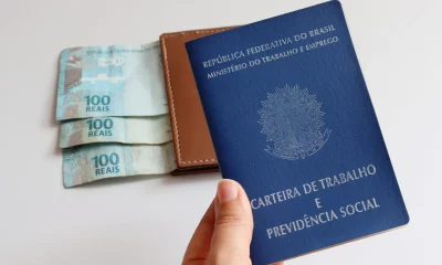 mão de um homem segurando a carteira de trabalho, com uma carteira com notas de 100 reais ao fundo