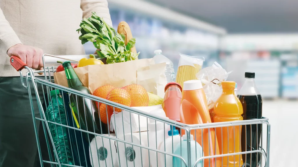 Carrinho de supermercado cheio com variedade de produtos, incluindo garrafas, sucos, papel higiênico, vegetais frescos, espaguete e laranjas, em um corredor de supermercado iluminado.