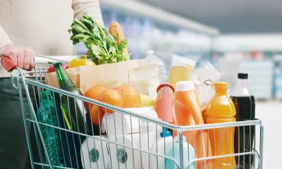 Carrinho de supermercado cheio com variedade de produtos, incluindo garrafas, sucos, papel higiênico, vegetais frescos, espaguete e laranjas, em um corredor de supermercado iluminado.